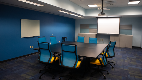 Conference room with large square table surrounded by chairs and projection screens on the wall.